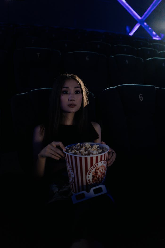 A woman sits in a dark theater holding a bucket of popcorn, immersed in a movie.